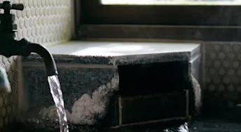 vattenskada-försäkring.webp Vattenkran med vatten som rinner ut i ett fuktskadat badkar byggt av blått och beige kakel.