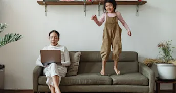insurance company En mamma jobbar vid datorn i en soffa medan ett barn hoppar i soffan.