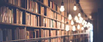 En upplyst hylla fylld av böcker En lång bokhylla fylld med böcker i dämpade färger, belyst av varma, hängande glödlampor som skapar en mysig atmosfär. Bakgrunden blir suddigare längre bort, vilket ger ett intryck av djup.