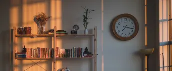 Klocka med träram på en vit vägg, bredvid en bokhylla full med böcker i olika färger