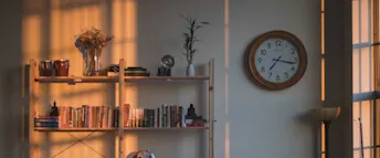 Klocka på vägg Klocka med träram på en vit vägg, bredvid en bokhylla full med böcker i olika färger