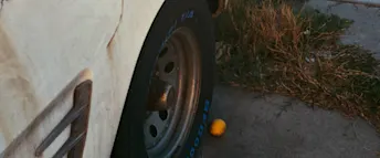 Närbild på ett bilhjul intill en gul frukt på marken, omgiven av vissna grässtrån och sliten asfalt. Slitet bildäck mot trottoarkant – vardagsmiljö med oväntad detalj.