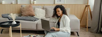 Din ränteguide 2024 Kvinna med mörkt hår som sitter på golv, lutandes mot grå soffa och läser på dator.