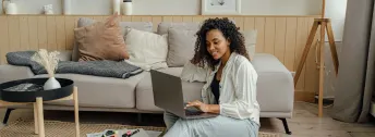 Kvinna med mörkt hår som sitter på golv, lutandes mot grå soffa och läser på dator.