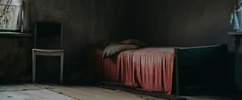 Mörkt och ödsligt rum med en obäddad säng och en tom stol bredvid ett fönster. En dunkel och övergiven interiör med en gammal trästol och en obäddad säng täckt av ett dammigt rosa täcke. Ljuset faller svagt in genom ett smutsigt fönster med trasiga gardiner, vilket förstärker känslan av ensamhet och övergivenhet i rummet.