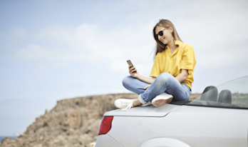 Kvinna sitter på en bil och kollar i mobilen