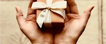 bröllopsgåva.webp två händer som håller i en liten ask med ett band som knutits till en rosett