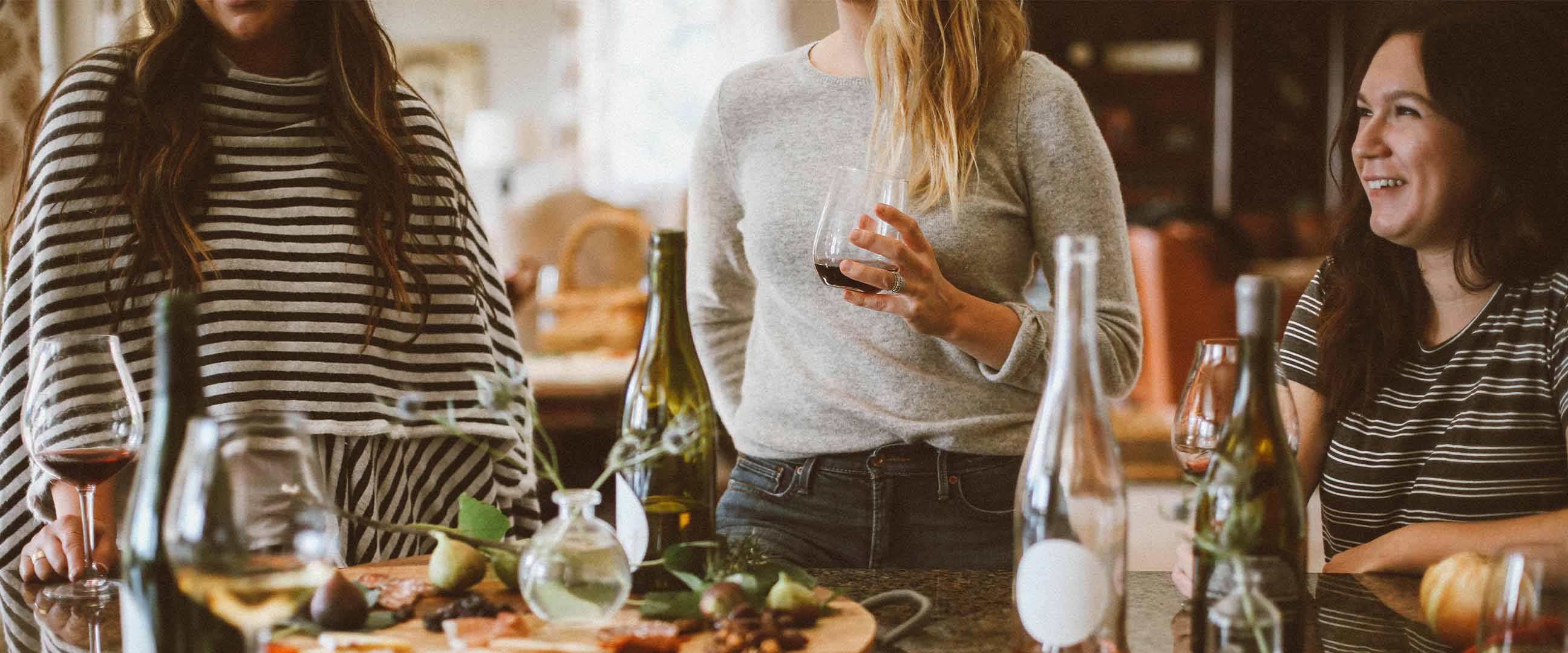 Tre vänner på restaurang – två står och en sitter framför ett bord med vinflaskor, glas och chark.