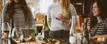 vänner-på-restaurang.jpg Tre vänner på restaurang – två står och en sitter framför ett bord med vinflaskor, glas och chark.