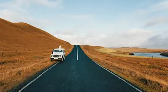 Starta bil med startkablar.webp Bil stående på sidan av en asfalterad väg, omgiven av torkade fält och en å.