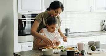Mor och dotter i kök Mor och dotter lagar mat i köket med vitt kakel och vita skåpsluckor. På köksbänken ligger det avokado och andra grönsaker.