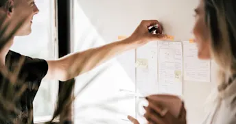 olika anställningsformer Kvinna sätter fast en lapp på en anslagstavla medan en kvinna med kaffekopp tittar på.