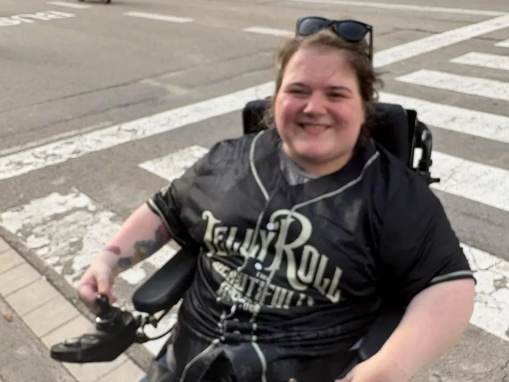 Niamh, a woman in a powered wheelchair, smiles happily while seated on the street.