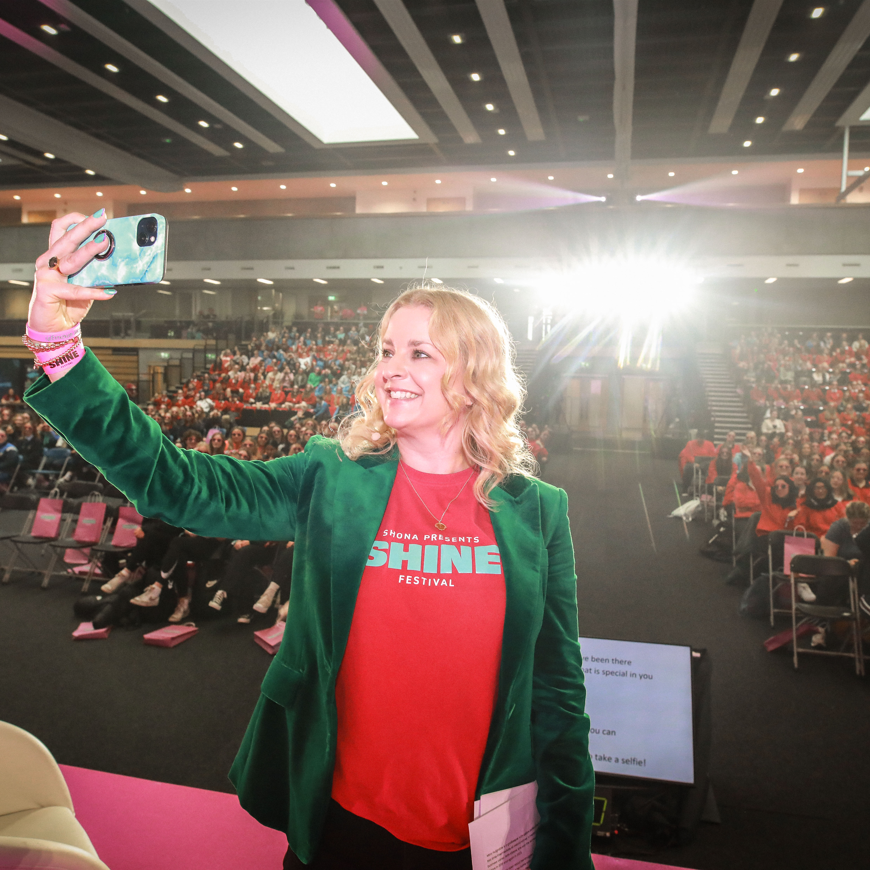 Tammy Darcy, a woman with a light skin tone, takes a selfie in front of a large audience with a range of light to dark skin tones. Many are wearing shirts that read, “The Shona Project Shine Festival.”
