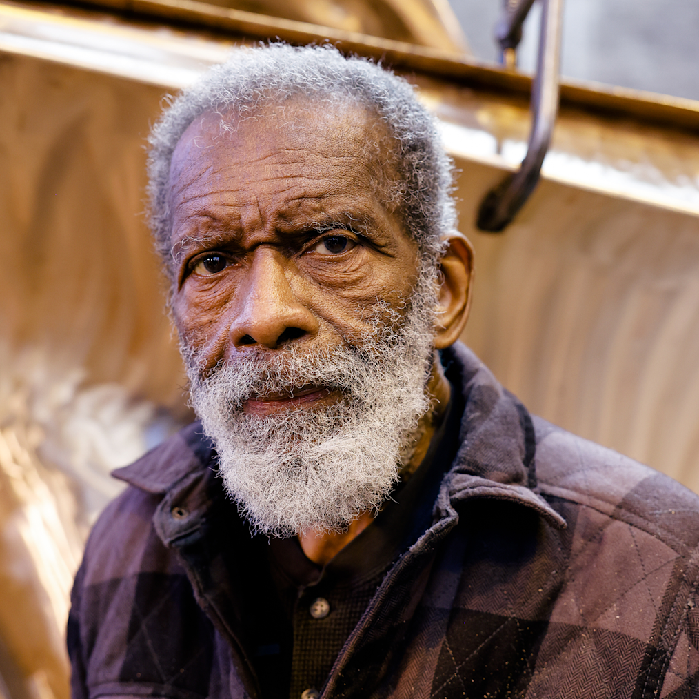 Richard Hunt, a Black man with a dark skin tone and gray hair and beard. He is not smiling. Behind him is a sculpture.