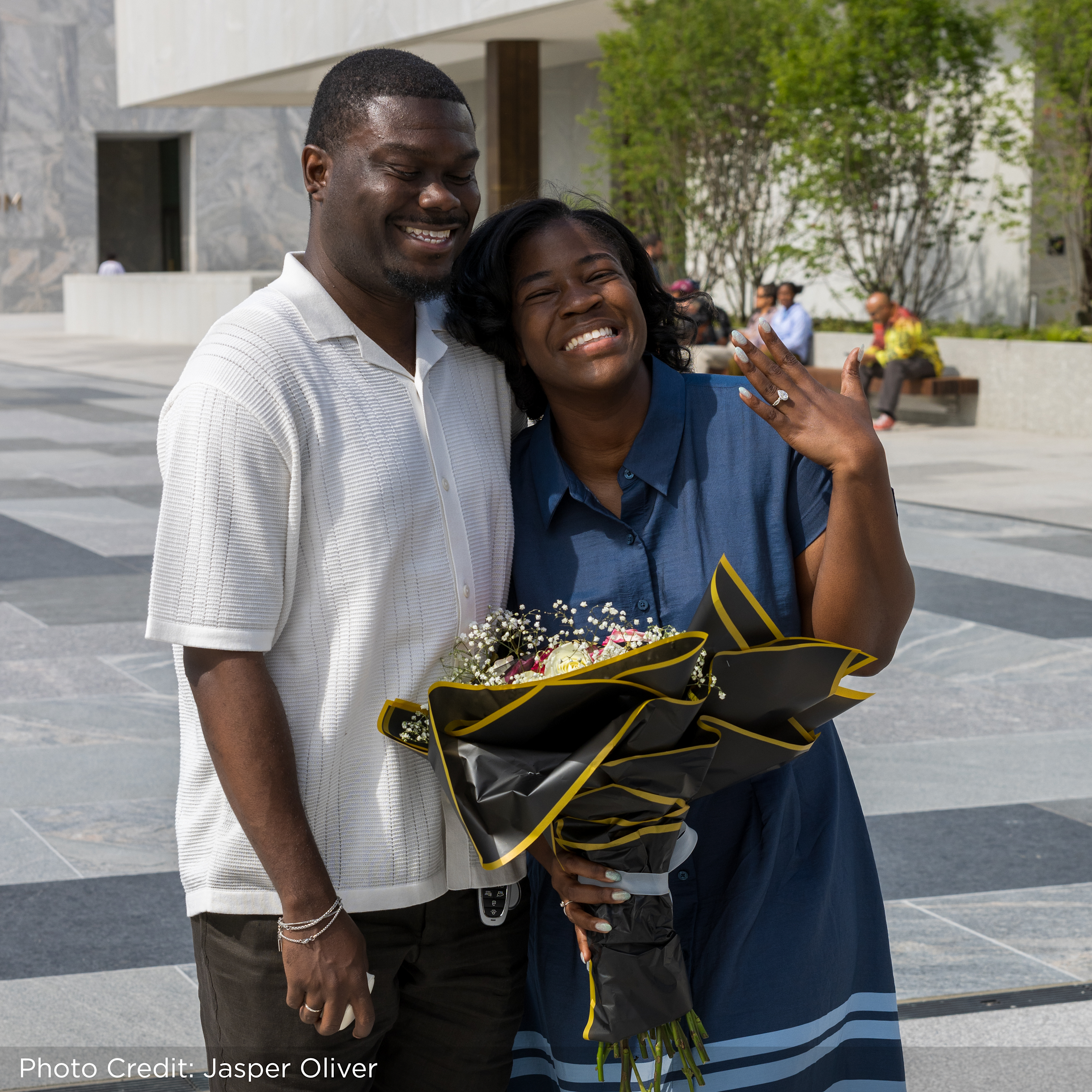 Vannessa Beene, a Black woman with a dark skin tone smiles as she holds up her left hand. She is posed next to her fiancé, Purvis Funches, a Black man with a dark skin tone. They are smiling as they stand on the John Lewis Plaza at the Obama Presidential Center.