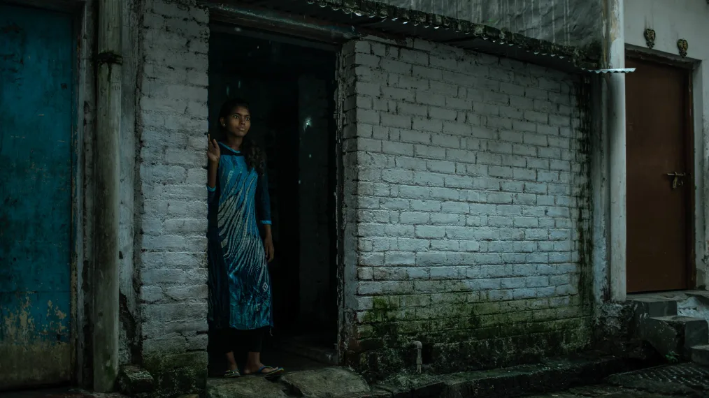 A girl with long hair with curly ends, deep skin, dot shaped earings, and a mostly blue, with black, and feint yellow accent dress stands holding the side of a door way. She looks out towards a road. The lighting is dark. The buildings are made of brick that has moss growing at the bottoms and flaking paint.