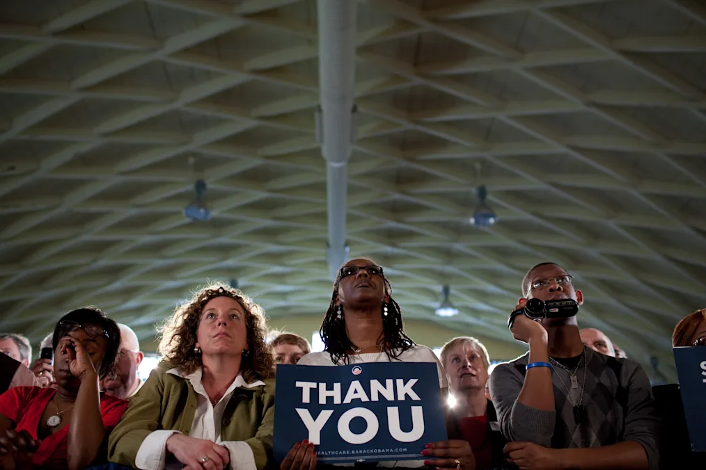 A woman with a deep skin tone wearing a white sweater holding a blue sign that says "thank you". There is a crowd of people of a variety of skin tones blurred out surrounding her. The ceiling is beige with a diamond pattern. 
