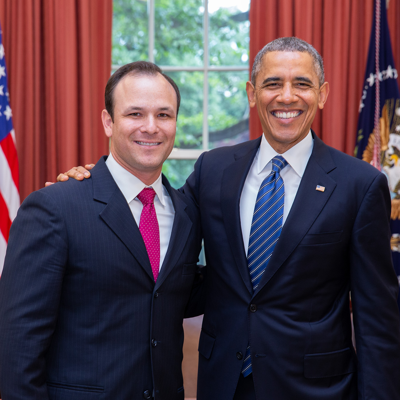A posed photo of Charlie Galbraith standing with President Barack Obama in the Oval Office in the White House. Behind them are the deep red Oval Office curtains and two flags: the American flag and the Flag of the President. President Obama has his right arm around Charlie’s shoulder. Both men are smiling in the photo. Charlie has an olive complexion and short dark hair. He is wearing a dark colored suit and a pink tie with a white shirt underneath. President Obama has a medium deep complexion and closely cut black and gray hair. He is wearing a dark suit and a blue striped tie with a white shirt underneath.