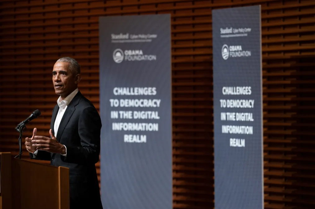 President Obama speaks into a microphone on the left of the image, with gray banners behind him that read "Challenges to Democracy in the Digital Realm"