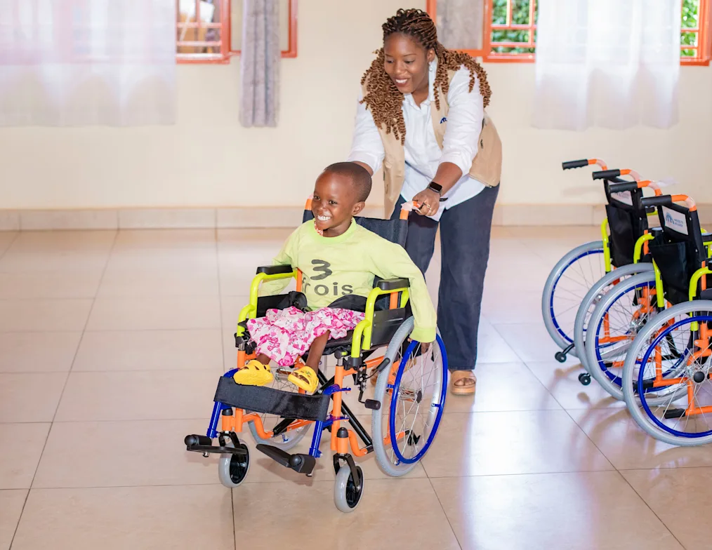 Arlette Irankunda, a Black woman with a dark skin tone and blonde braids, smiles as she pushes a smiling patient in a wheelchair.