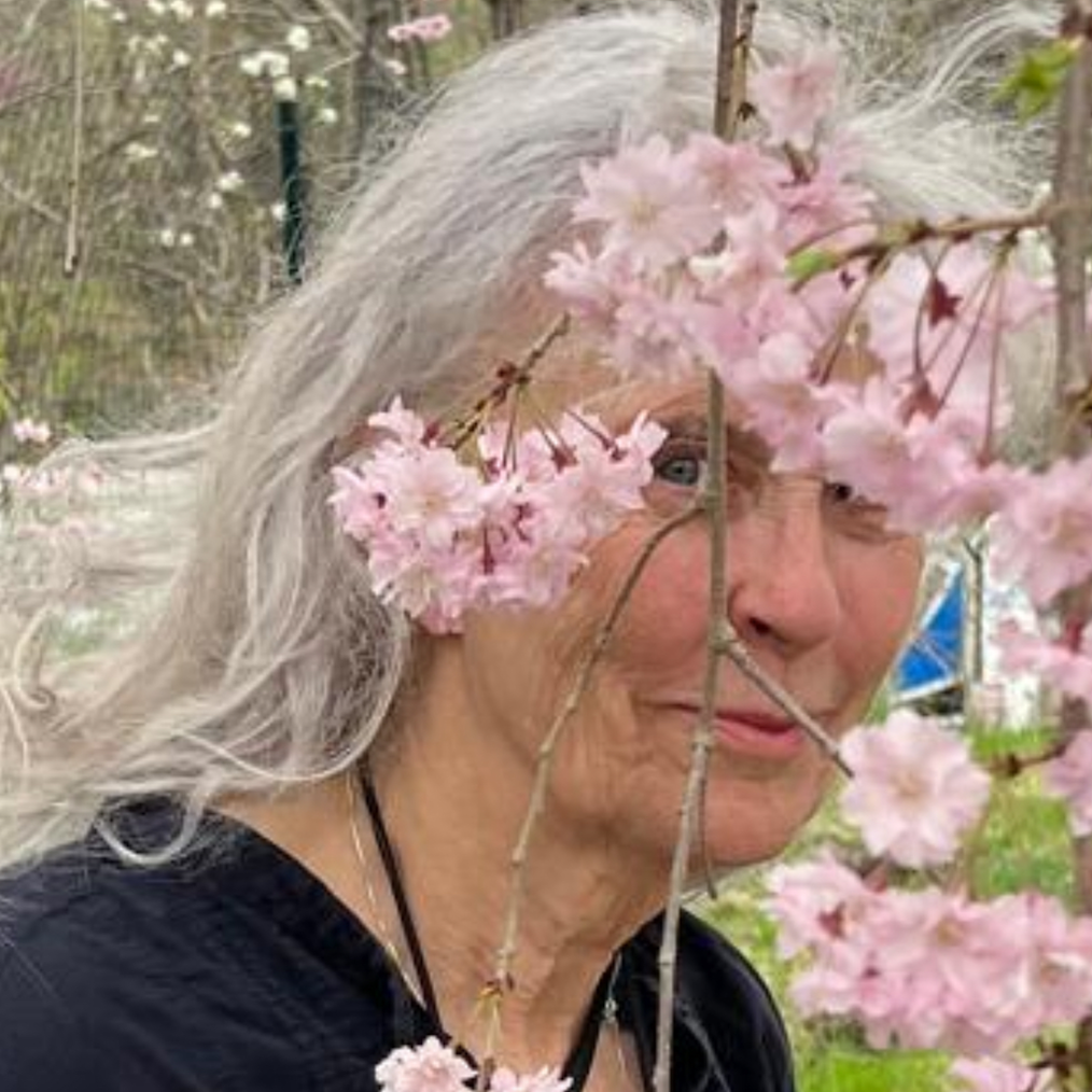 A photographed portrait of Kiki Smith. Kiki has a light olive complexion and long gray hair. She is standing outside in a field, her face is partially obscured by pink flowers hanging from the right side of the image. She is wearing a black shirt.