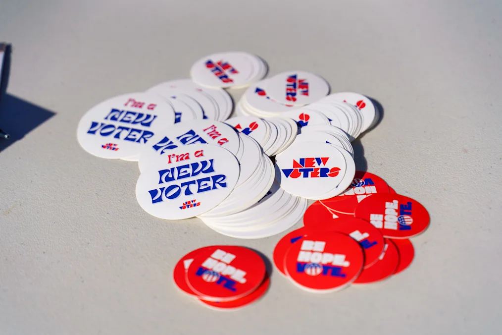 A pile of round stickers sits scattered on an off-white table surface. Several white stickers feature blue and red letters reading "I'm a NEW VOTER" or "NEW VOTERS". A cluster of smaller red circular stickers reads "BE HOPE. VOTE." in bold white and blue text.