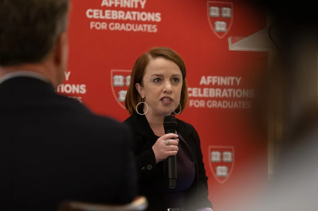 Rebecca speaks at Harvard Kennedy School affinity celebration for disabled graduates. She is wearing silver hoop earrings, a purple dress, and a black blazer. She is sitting in front of a red background that reads, “Affinity Celebration for Graduates.”