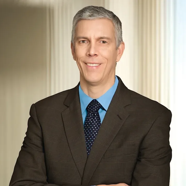 A waist-up headshot of a White man with gray hair, blue shirt and tie and dark suit jacket. His arms are crossed and he smiles slightly, showing teeth.