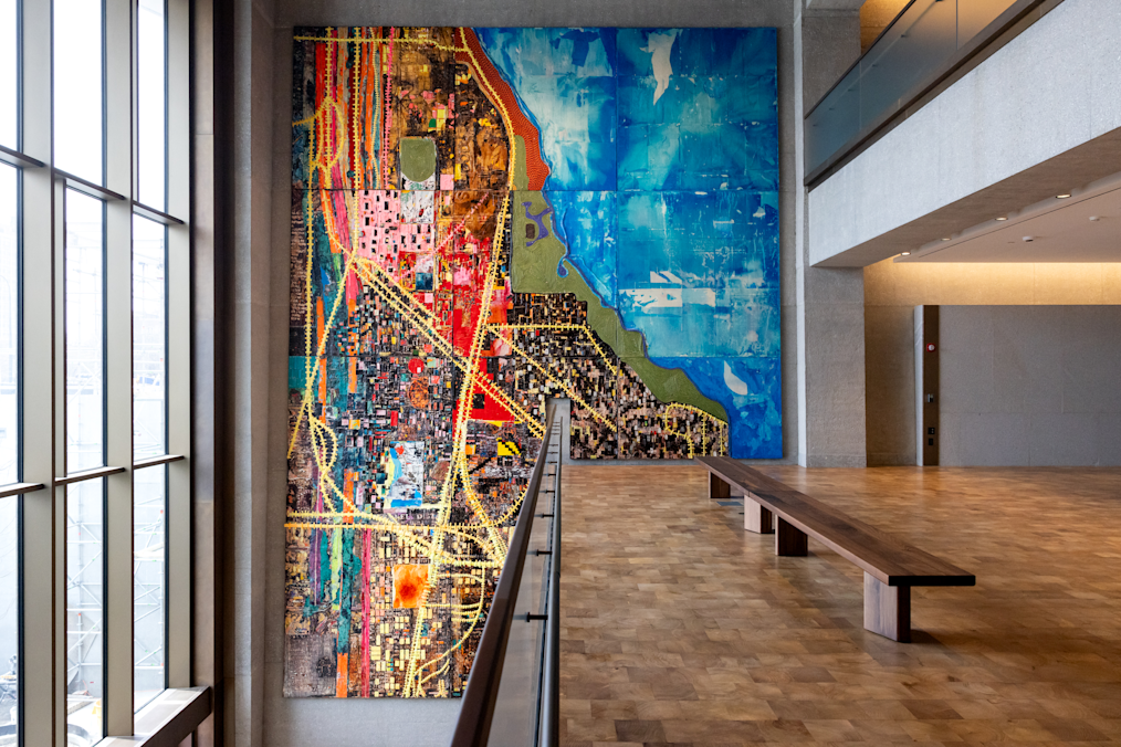 A high-angle photo shows a spacious, upper-level walkway overlooking an atrium with parquet wood flooring and smooth concrete walls. On the right, a long wooden bench sits parallel to the edge of the floor. In the center back, a tall, vertical tapestry hangs on the wall, featuring grid-like, abstract patterns with yellow, pink, red, black, and bright blue colors reminiscent of a colorful city map. The tapestry is called the City of the Big Shoulders, a new installation from Mark Bradford.