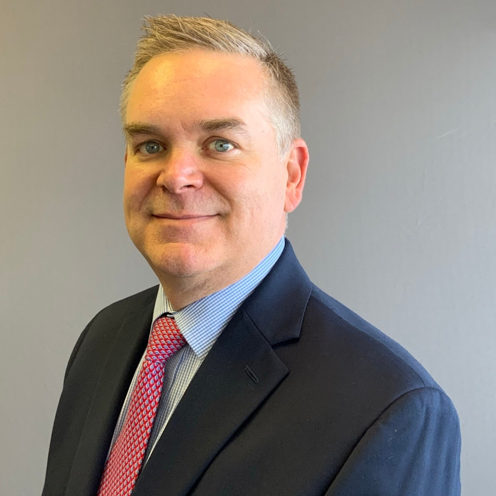 Joe Skeel, a man with a light skin tone and blonde hair, holds a closed lipped smile. He is wearing a navy suit, blue shirt, and red tie.
