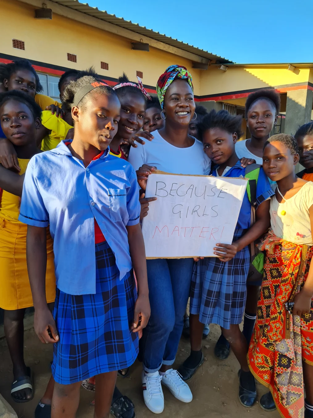 Ireen Chikatula poses for a photo with 11 teenage students. They have a deep skin tone and some are wearing school uniforms. Ireen Chikatula holds a sign that reads, “Because girls matter!”