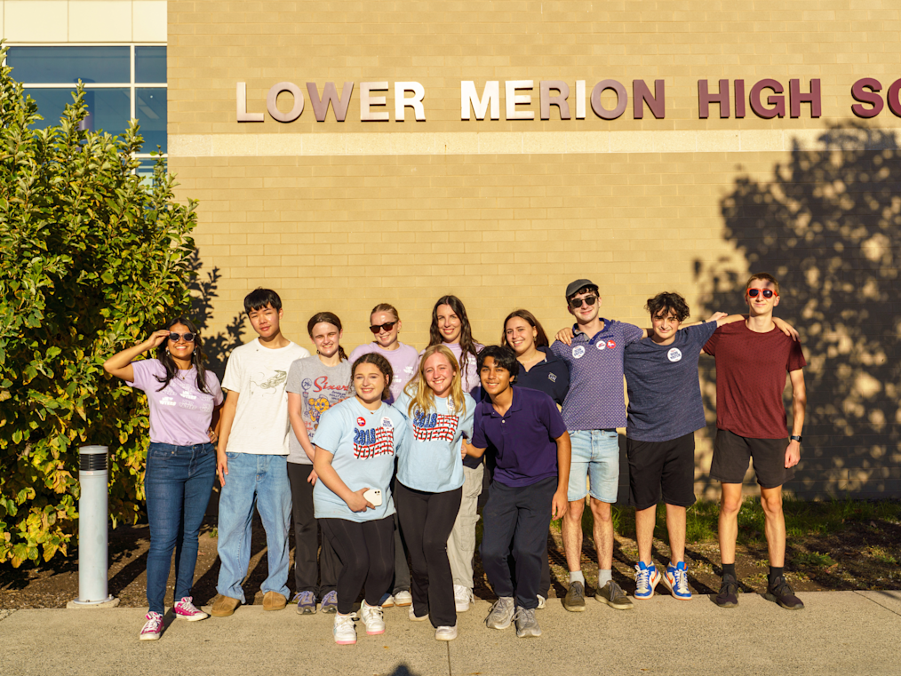 Ten smiling young people stand shoulder-to-shoulder on a sidewalk in front of a brick high school building. They have light, medium-light, and medium skin tones. Above them, large brown lettering on the beige brick wall reads "LOWER MERION HIGH SCHOOL". The scene is bathed in warm afternoon sunlight.