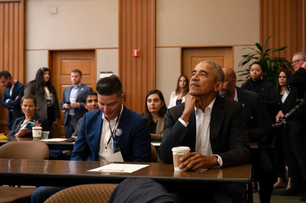 President Obama and other people of various skin tones sit and stand as they are interacting and listening