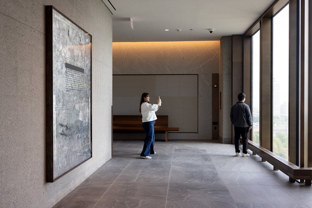 A woman takes a photo of a large artwork mounted on a concrete wall inside a sunlit hallway next to floor-to-ceiling windows. The room is the Sky Room Vista