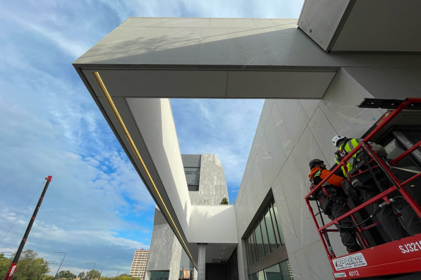 The photograph is a look at the lighting fixture above one of the courtyards on the Obama Presidential Center campus. The picture is taken from below. The lighting fixture is long and rectangular with one long singular light. Next to the lighting fixture is a red crane with two construction workers.