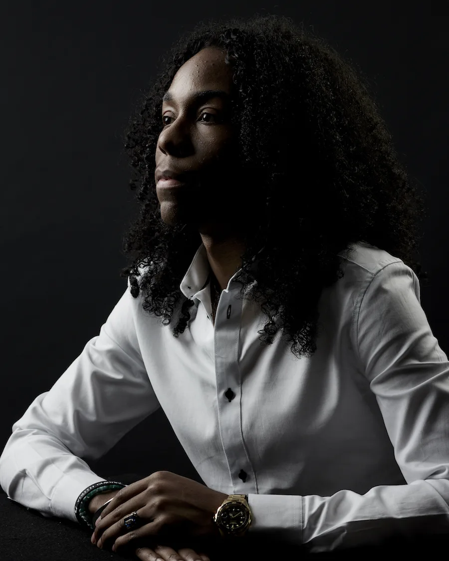 Christian Champagne, a Black man with a deep skin tone looks off camera. He has long black hair and is wearing a white shirt. 