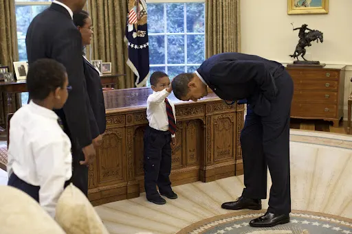 A young Black boy reaches out to touch President Obama's hair. They are in the oval office and a two other adults and one child stand to the side.