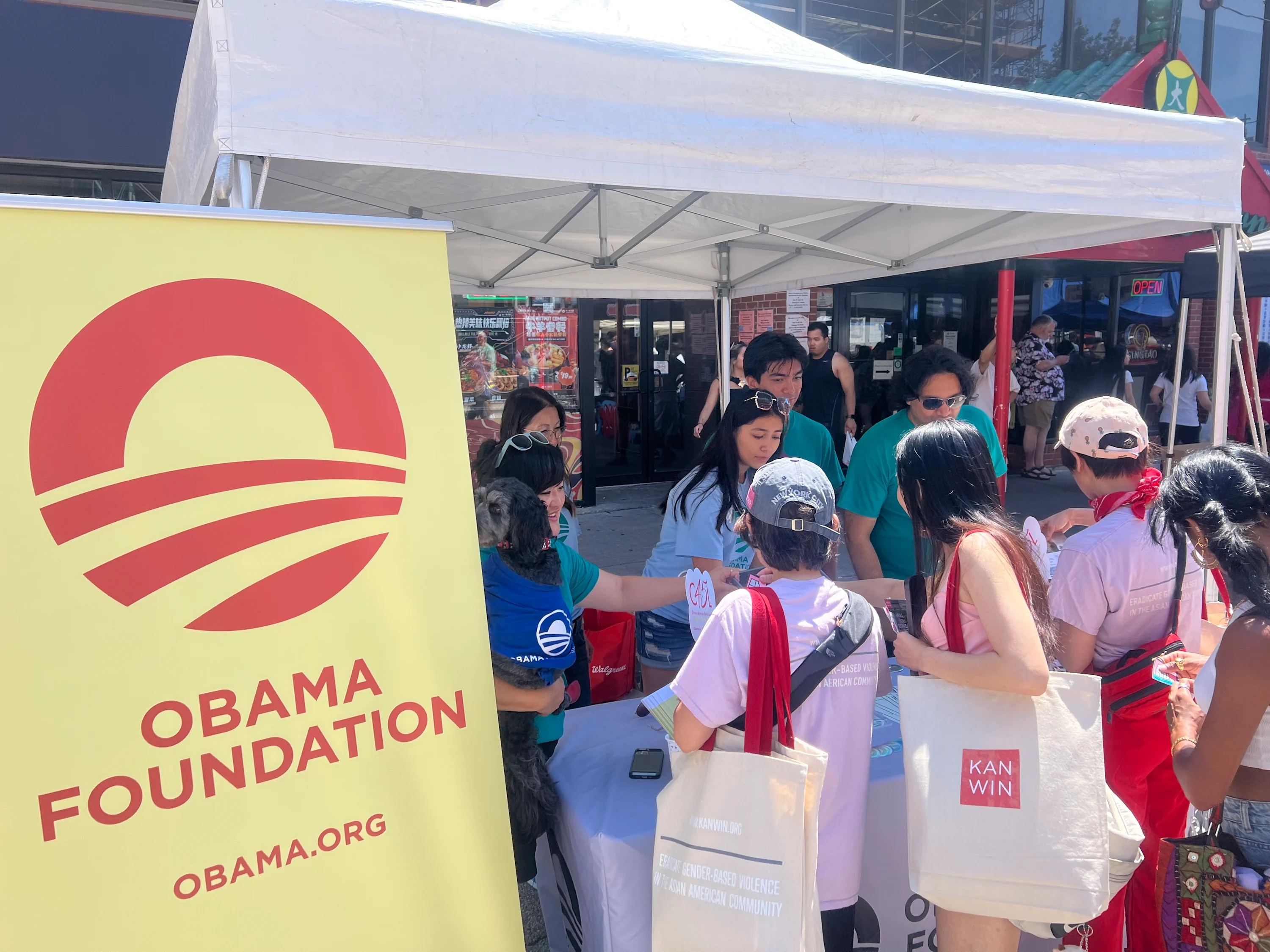 A yellow Obama Foundation banner with a red logo is seen beside a table. The banner reads, “Obama Foundation” and “Obama.org.” At the table, a group of people with light to medium skin tones interact with one another at a street festival.