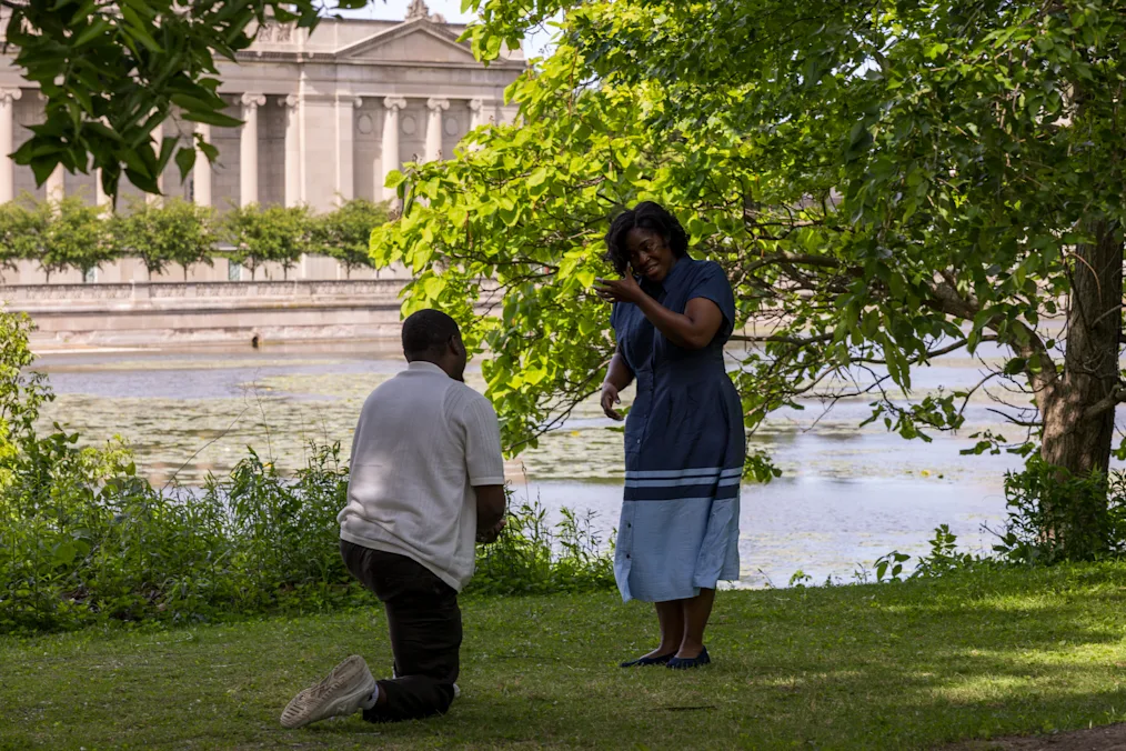A shot of Purvis Funches, a Black man with a dark skin tone and low black hair, on one knee as he proposes to Vannessa Beene, a Black woman with a dark skin tone. She is wearing a blue dress.