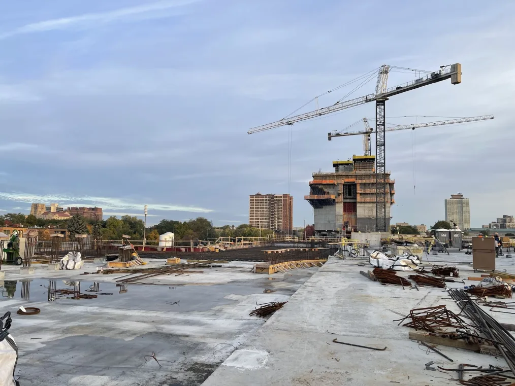 A view of the Museum Building rising to the sky on the site of the Obama Presidential Center. Cranes are in the sky as work continues on the garage rooftop. Construction equipment is scattered throughout.