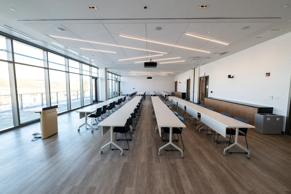 A look at a multipurpose room in Home Court. The room features several long white tables with several dark colored chairs. In the front of the room is a light-colored wooden podium. Behind the podium are several windows.