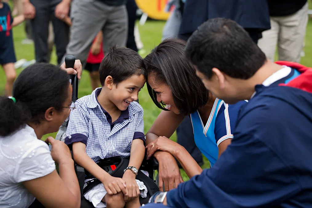 First Lady Michelle Obama sits head to head with a young boy with a light-medium skin tone, a blue and white plaid polo shirt sitting in a wheelchair. There are two individuals with a light-medium and medium skin tones squatting in front of First Lady Michelle Obama and the young boy. 