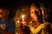 Barbara Mukisa, a Black girl with a dark skin tone, holds a candle as she smiles at the camera. She has low hair and is wearing a colorful patterned top. Girls in the background are slightly blurred.