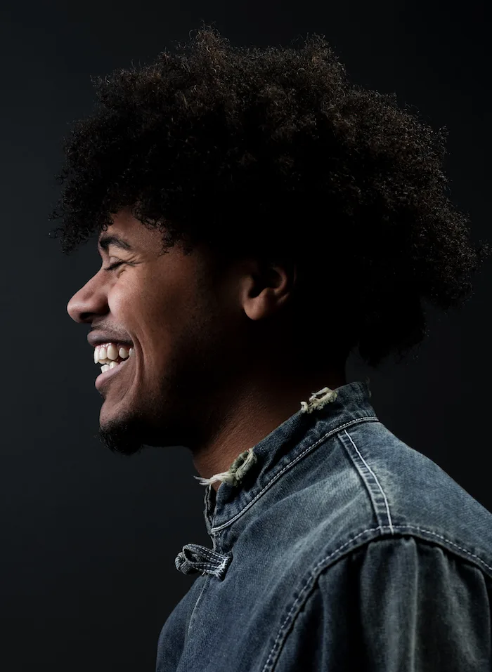 A side profile portrait photo of a man with a medium skin tone wearing a denim jacket looking toward the left-hand side of the photo and smiling in front of a gradient black background. 