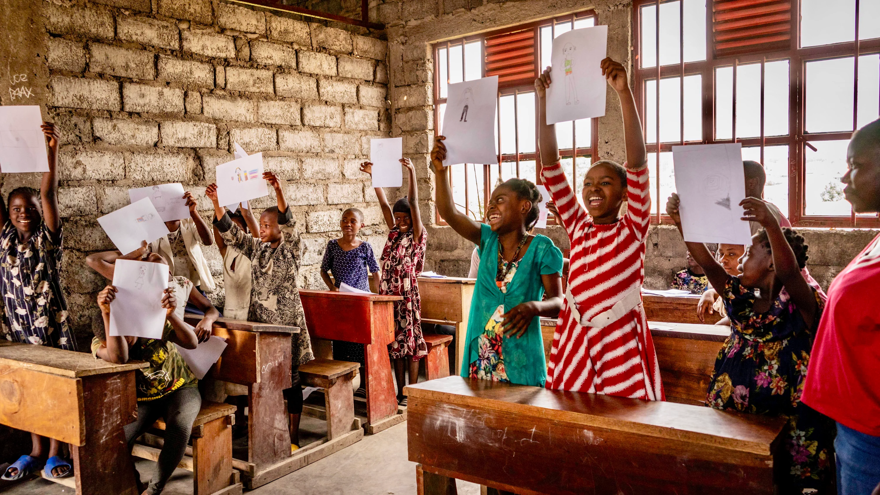 A group of young girls with a range of medium and dark skin tones hold up sheets of paper in a classroom. All girls are standing behind wooden desks. 