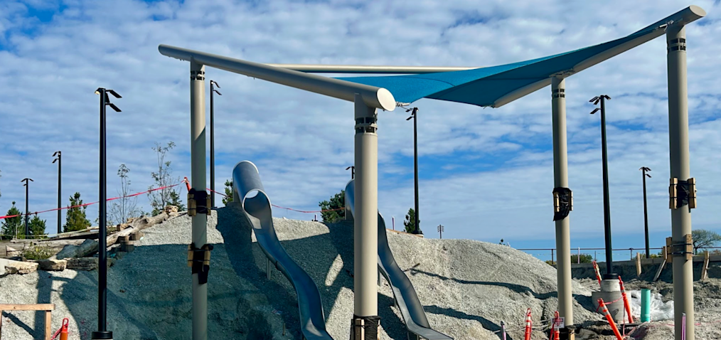 A photograph of the Obama Presidential Center Playground. The image features an outdoor canopy and two silver metal slides that stretch down a hill. In the background is a large dirt mound.