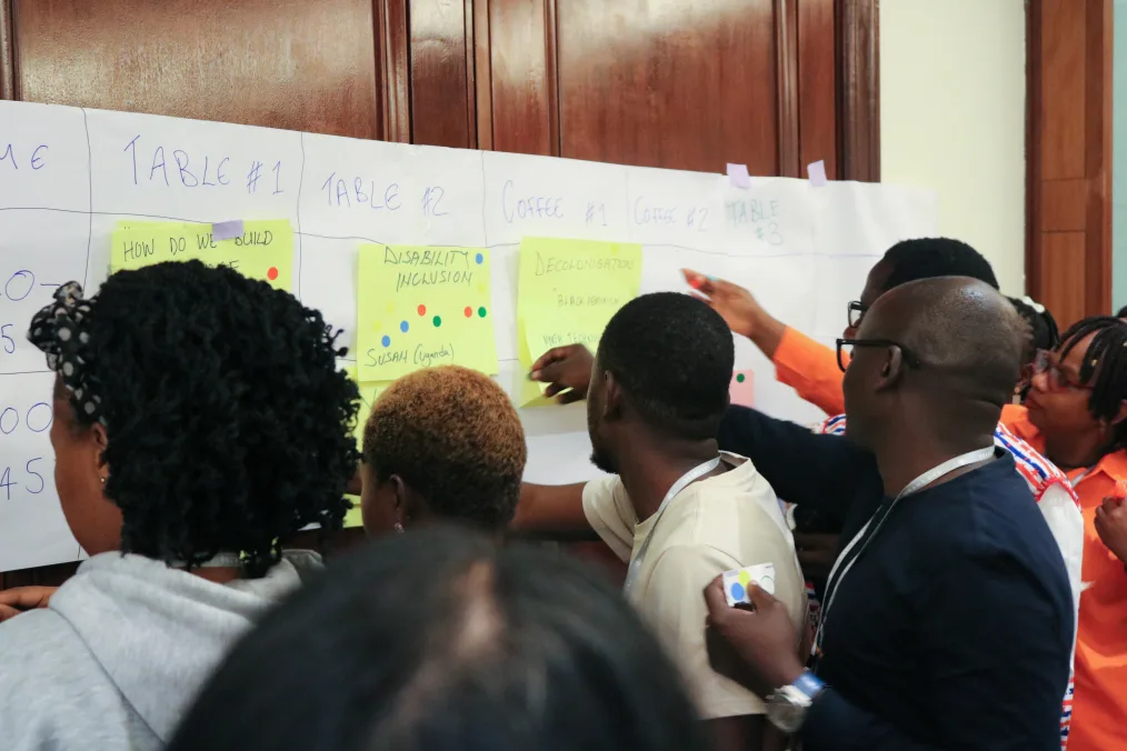 A group of people with a range of medium and deep skin tones place stickers on post it notes on a wall in a room. Their backs are to the camera. The post it notes read, “disability inclusion” and “decolonisation.”