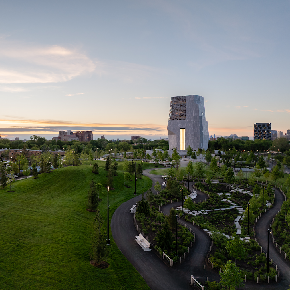 A high-angle view shows a green lawn and winding dark paths next to a tall stone building at dusk. Small trees and plants line the paths under a glowing sky.