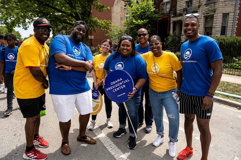Seven Obama Foundation employees, with a range of light to dark skin tones, pose for a photo with community members at a street festival.
